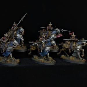 May include: Miniature figurines of mounted knights in armor, each holding a lance. The knights wear blue and silver armor with red accents. The horses are brown and tan, and the bases are oval with a grassy texture.
