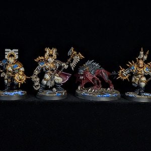 Puede incluir: Cuatro figuras de fantasía en miniatura. Dos figuras son guerreros con armadura, hachas, detalles dorados y azules. Un monstruo rojo y negro con dientes y garras afiladas se interpone entre ellos. La cuarta figura es otro guerrero con un hacha.