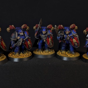 Może przedstawiać: Grupa sześciu pomalowanych na niebiesko i czerwono figurek z mieczami i tarczami. Figurki stoją na okrągłych podstawach.