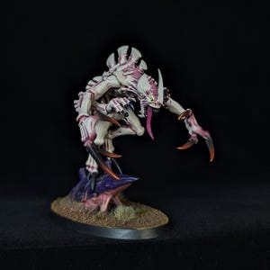 Puede incluir: Figura en miniatura de una criatura blanca y morada, con garras afiladas y una larga lengua. Se encuentra sobre una base texturizada. El fondo es negro.