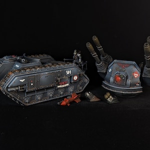 Könnte beinhalten: Detailliertes Miniaturmodell eines dunkelgrauen Panzers mit der Nummer 384 an der Seite, zusammen mit einem separaten Geschützturm und mehreren kleinen Raketen. Der Panzer weist komplizierte Details und ein rotes Emblem auf. Die Szene ist vor schwarzem Hintergrund.