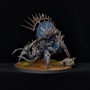 May include: A detailed miniature figure of a mechanical creature with a blue and gold color scheme. The figure has multiple legs, sharp claws, and a spiked crown. It is set on a brown base with small patches of green grass.