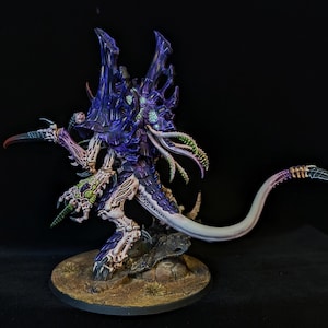 May include: A detailed miniature figure of a creature with a purple and blue exoskeleton, sharp claws, and multiple appendages. The figure is mounted on a textured, circular base, showcasing intricate details and vibrant paintwork.