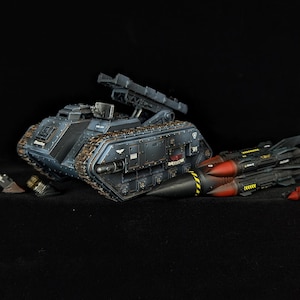 Könnte beinhalten: Detailliertes Modell eines dunkelblauen Militärpanzers mit Raketenwerfer und mehreren Raketen. Der Panzer hat eine strukturierte Oberfläche und wird von roten und schwarzen Raketen mit gelben Streifen begleitet.