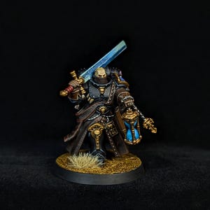以下が含まれることがあります： 黒い鎧に金の縁取りを施した戦士のミニチュアフィギュア。肩には光る青い剣を担ぎ、もう一方の手には青いランタンを持っている。長いダークコートを着用し、草が生えたテクスチャーのある台座の上に立っている。