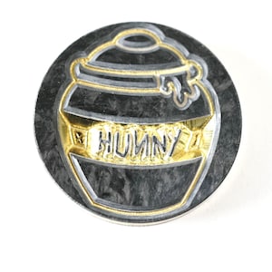 Op de afbeelding: Een ronde, zwarte en gouden metalen pin met een honingpotontwerp. De honingpot is gegraveerd met het woord "HONEY" in gouden letters.