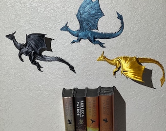 May include: Three dragon ornaments in black, blue, and gold are positioned above four books. The books are titled "Fourth Wing" and "Iron Flame" by Rebecca Yarros. The dragons are depicted in flight.