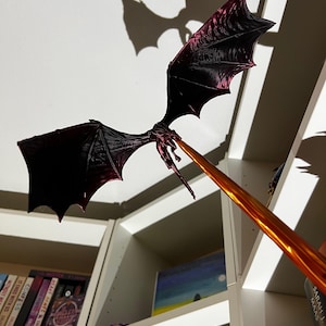 May include: A black and purple dragon figurine with outstretched wings, mounted on a gold-colored stick. The dragon is positioned against a white wall with a bookshelf in the background.