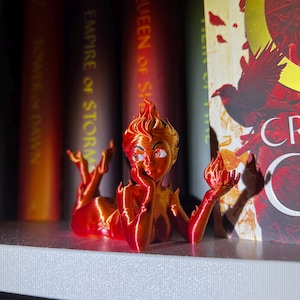 May include: A 3D-printed figurine of a reclining woman with fiery hair and flames. The figure is primarily red and orange, with details in a lighter orange. The figurine is resting on a white shelf in front of a row of books.