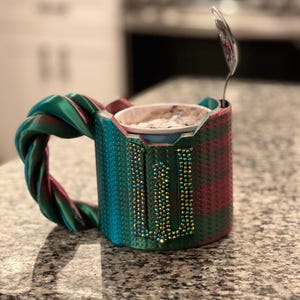 Puede incluir: Una taza turquesa, roja y verde con un asa trenzada. La taza tiene una letra "U" enjoyada en la parte delantera. Una cuchara pequeña descansa dentro de la taza, que contiene una sustancia blanca. La taza está sobre una encimera de granito.