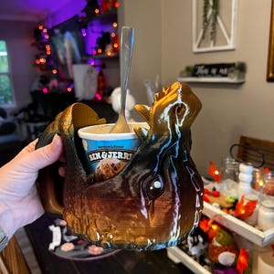 May include: A brown and black decorative mug with a unique, textured design, holding a container of Ben & Jerry's ice cream. A silver spoon rests inside the ice cream. The mug has a handle and is held by a person.