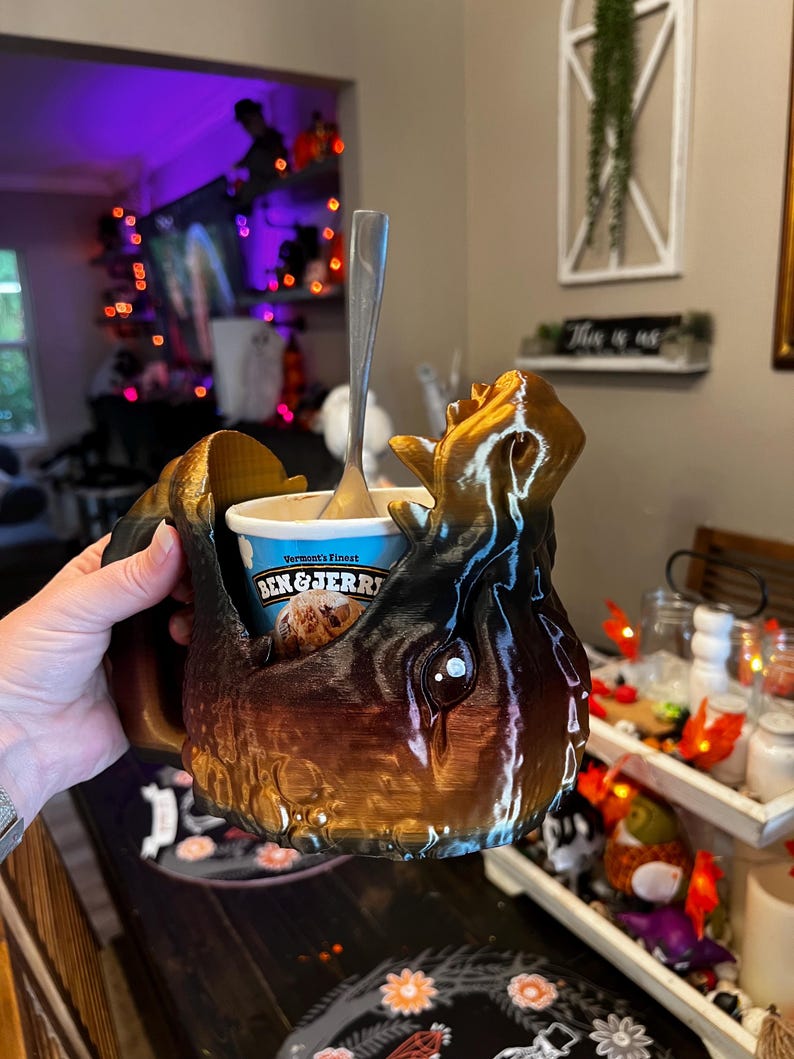 May include: A brown and black decorative mug with a unique, textured design, holding a container of Ben & Jerry's ice cream. A silver spoon rests inside the ice cream. The mug has a handle and is held by a person.