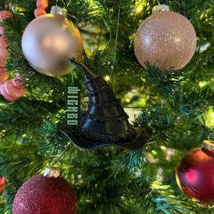 May include: A black witch hat ornament with the word "Wicked" in green lettering hangs on a Christmas tree. The tree is decorated with gold and red ornaments.