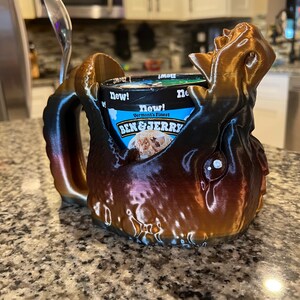 May include: A 3D-printed ice cream holder in shades of brown and bronze, designed to resemble a creature. The holder cradles a pint of Ben & Jerry's ice cream, with a spoon resting on the side. The words "New!" and "Vermont's Finest" are visible.