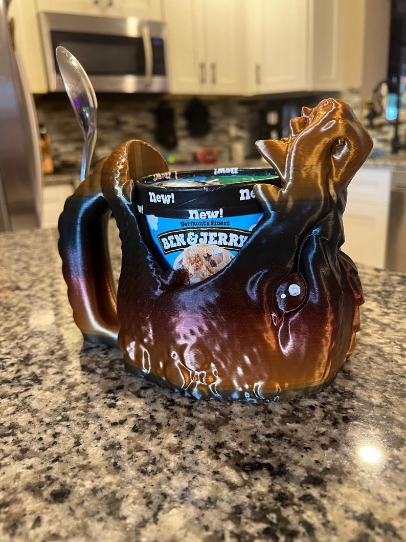 May include: A 3D-printed ice cream holder in shades of brown and bronze, designed to resemble a creature. The holder cradles a pint of Ben & Jerry's ice cream, with a spoon resting on the side. The words "New!" and "Vermont's Finest" are visible.