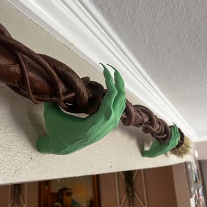 Może przedstawiać: Dekoracja na Halloween przedstawiająca brązową, skręconą winorośl trzymaną przez zielone, przypominające pazury dłonie. Winorośl rozciąga się na białej powierzchni. Projekt ma upiorny, fantastyczny motyw.