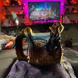 May include: A brown and black decorative mug with a handle, holding a pint of Ben & Jerry's ice cream. A silver spoon rests on the rim. The mug has a textured, ornate design. The background shows a television and Halloween decorations.