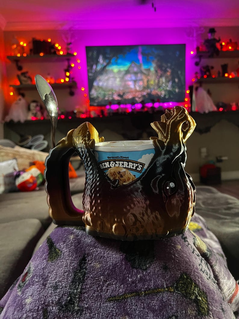 May include: A brown and black decorative mug with a handle, holding a pint of Ben & Jerry's ice cream. A silver spoon rests on the rim. The mug has a textured, ornate design. The background shows a television and Halloween decorations.