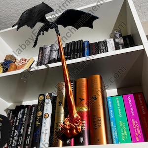 May include: A black dragon figurine with outstretched wings, perched on a bookshelf with a variety of fantasy novels. The dragon's tail is a fiery orange colour and is pointing upwards.