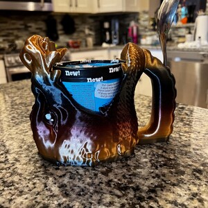May include: A 3D-printed ice cream pint holder in brown, black, and gold tones, with a handle and a slot for a spoon. The holder is designed to cradle a pint of ice cream, with the word "new!" visible on the container.