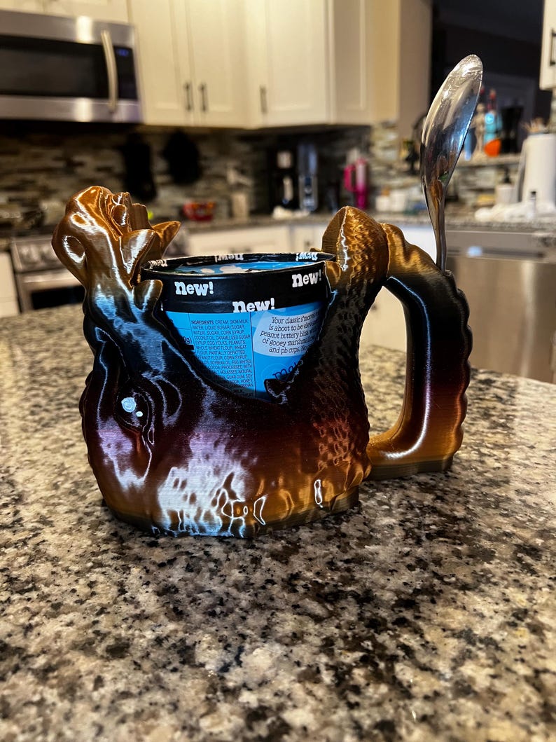 May include: A 3D-printed ice cream pint holder in brown, black, and gold tones, with a handle and a slot for a spoon. The holder is designed to cradle a pint of ice cream, with the word "new!" visible on the container.