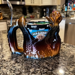 May include: A 3D-printed ice cream mug holder in shades of brown, black, and bronze, designed to cradle a pint of Ben & Jerry's ice cream. The mug has a unique, organic shape with a handle and a spoon.