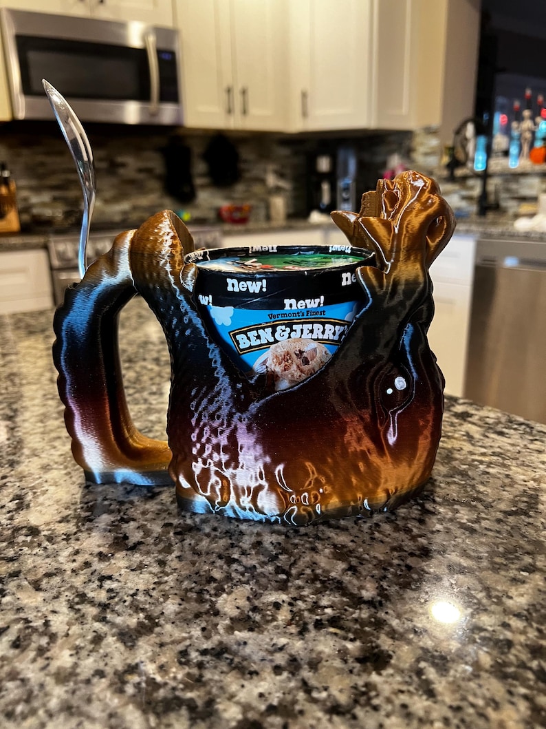 May include: A 3D-printed ice cream mug holder in shades of brown, black, and bronze, designed to cradle a pint of Ben & Jerry's ice cream. The mug has a unique, organic shape with a handle and a spoon.