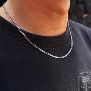 Könnte beinhalten: Eine silberne Panzerketten-Halskette auf einem schwarzen T-Shirt. Die Kette besteht aus kleinen, ineinandergreifenden Gliedern. Die Halskette hat ein schlichtes, klassisches Design. Das Bild ist gut beleuchtet und hebt die Details der Halskette hervor.