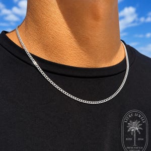 May include: A silver-toned curb chain necklace resting against a black shirt. The necklace features interlocking links with a polished finish. A circular logo in the bottom right corner displays a palm tree and the text AZURE JEWELS ESTD 2024.