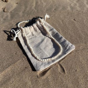 May include: A delicate pearl necklace rests on a beige drawstring pouch. The necklace is composed of small, round, white pearls. The pouch is made of a textured fabric and is lying on a sandy surface.