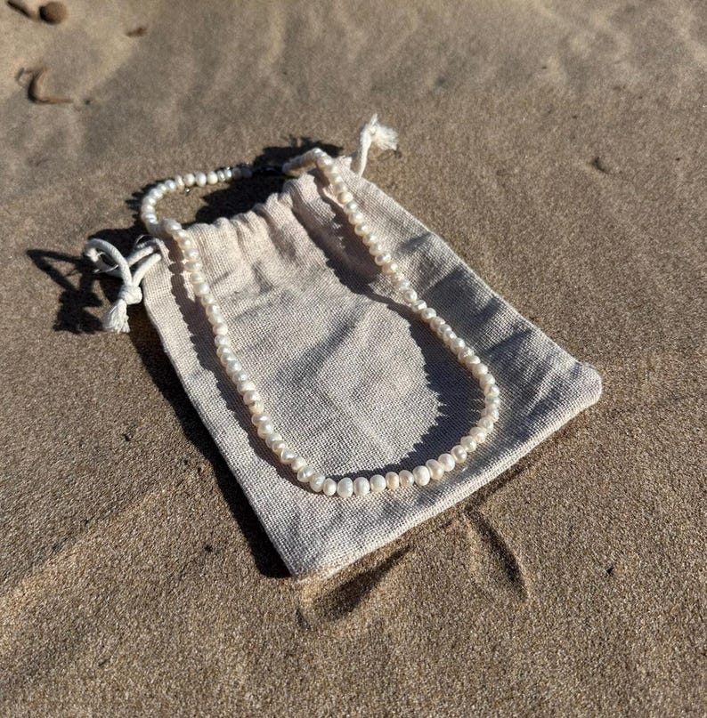 May include: A delicate pearl necklace rests on a beige drawstring pouch. The necklace is composed of small, round, white pearls. The pouch is made of a textured fabric and is lying on a sandy surface.
