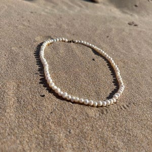 May include: A delicate pearl necklace resting on a sandy surface. The necklace features numerous small, round, white pearls strung together. The background is a light brown, sandy texture.
