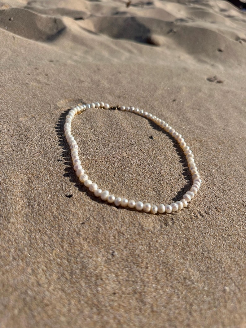 May include: A delicate pearl necklace resting on a sandy surface. The necklace features numerous small, round, white pearls strung together. The background is a light brown, sandy texture.