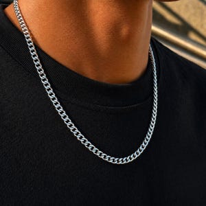 May include: A silver-toned metal chain necklace featuring a classic curb link design. The necklace is worn around the neck against a plain black t-shirt, showcasing the polished, reflective surface of the interlocking metal links.