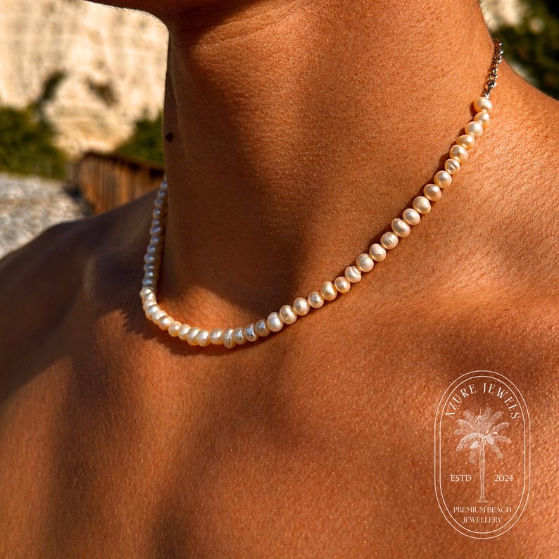 May include: A delicate pearl necklace with a silver clasp. The necklace is composed of small, round, creamy-white pearls. The image is of a person wearing the necklace. The necklace is a piece of jewelry.