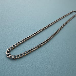 May include: A silver-toned metal chain necklace featuring a classic curb link design. The necklace is composed of interlocking, flattened, polished links that create a continuous, uniform pattern throughout the entire length of the jewelry piece.
