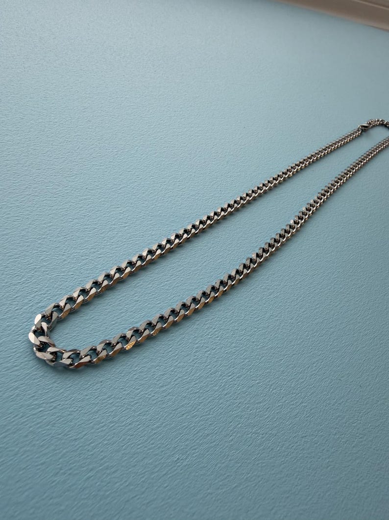 May include: A silver-toned metal chain necklace featuring a classic curb link design. The necklace is composed of interlocking, flattened, polished links that create a continuous, uniform pattern throughout the entire length of the jewelry piece.