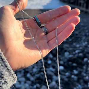 以下が含まれることがあります： 繊細なカーブリンクデザインのシルバーチェーンネックレス。2つの指輪が見え、1つはターコイズと赤のインレイ、もう1つは模様入りです。ジュエリーは手で持たれており、背景はぼやけた海です。