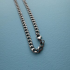 May include: A silver-toned metal chain necklace featuring interlocking flat curb links. The chain is constructed with a polished finish and includes a secure lobster claw clasp mechanism at one end, allowing for easy fastening and adjustment.
