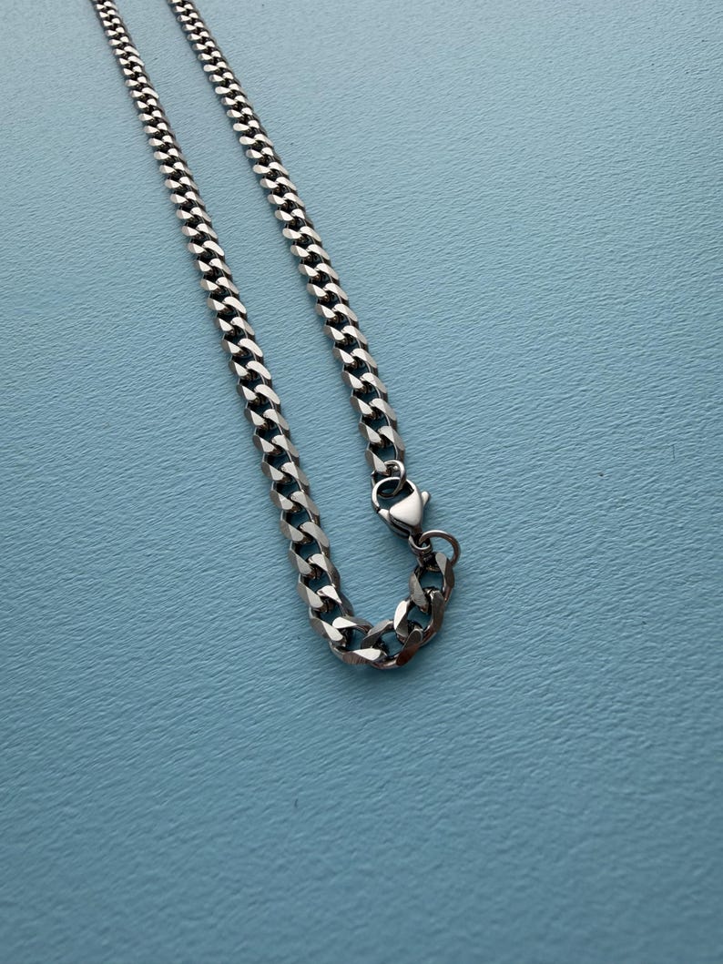 May include: A silver-toned metal chain necklace featuring interlocking flat curb links. The chain is constructed with a polished finish and includes a secure lobster claw clasp mechanism at one end, allowing for easy fastening and adjustment.