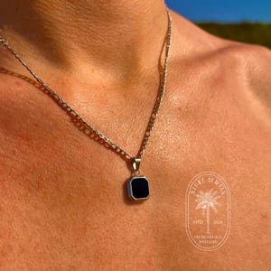 May include: A silver chain necklace with a square pendant featuring a black stone. The pendant has a beveled edge and a polished finish. The necklace is worn by a person with tanned skin.