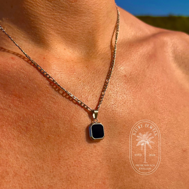 May include: A silver chain necklace with a square pendant featuring a black stone. The pendant has a beveled edge and a polished finish. The necklace is worn by a person with tanned skin.
