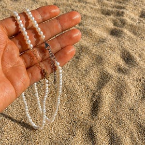May include: A delicate pearl necklace with a gold and silver clasp. The necklace is composed of small, round, white pearls. The background is a sandy beach, with the necklace held in a hand.