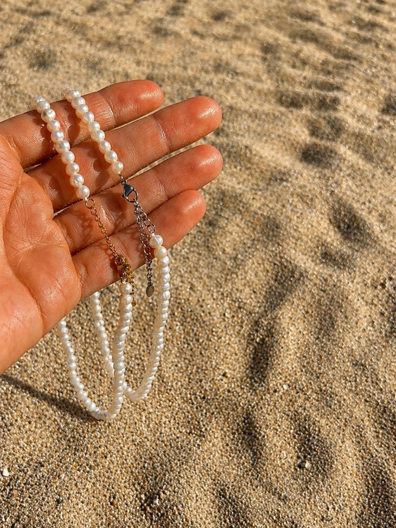 May include: A delicate pearl necklace with a gold and silver clasp. The necklace is composed of small, round, white pearls. The background is a sandy beach, with the necklace held in a hand.