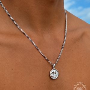 May include: A silver-toned curb chain necklace featuring a circular pendant with a raised wave design. The pendant has a polished metallic finish and is suspended from a simple metal bail. The necklace rests against the skin.