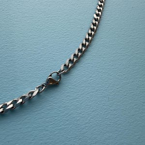 May include: A silver-toned metal chain necklace featuring interlocking flat curb links and a secure lobster claw clasp mechanism. The chain has a polished, reflective finish and is displayed in a curved arrangement on a solid light blue surface.