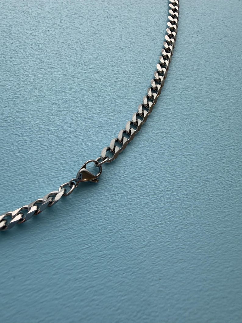 May include: A silver-toned metal chain necklace featuring interlocking flat curb links and a secure lobster claw clasp mechanism. The chain has a polished, reflective finish and is displayed in a curved arrangement on a solid light blue surface.