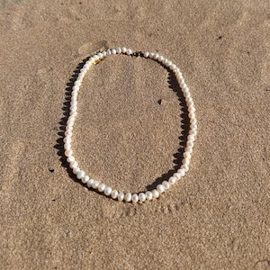 May include: A delicate pearl necklace resting on a sandy surface. The necklace features small, round, off-white pearls strung together. The clasp appears to be gold-toned. The necklace is centered in the frame, casting a soft shadow on the sand.