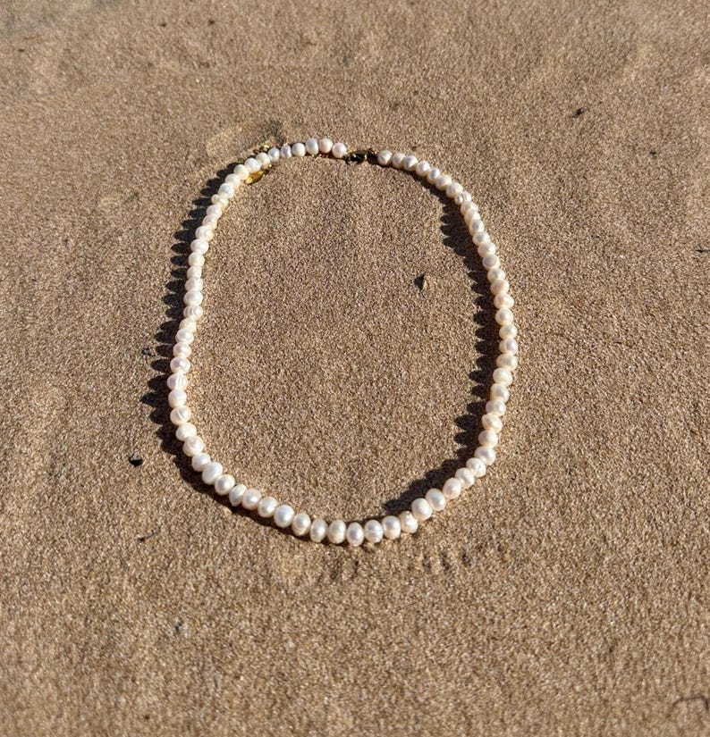 May include: A delicate pearl necklace resting on a sandy surface. The necklace features small, round, off-white pearls strung together. The clasp appears to be gold-toned. The necklace is centered in the frame, casting a soft shadow on the sand.