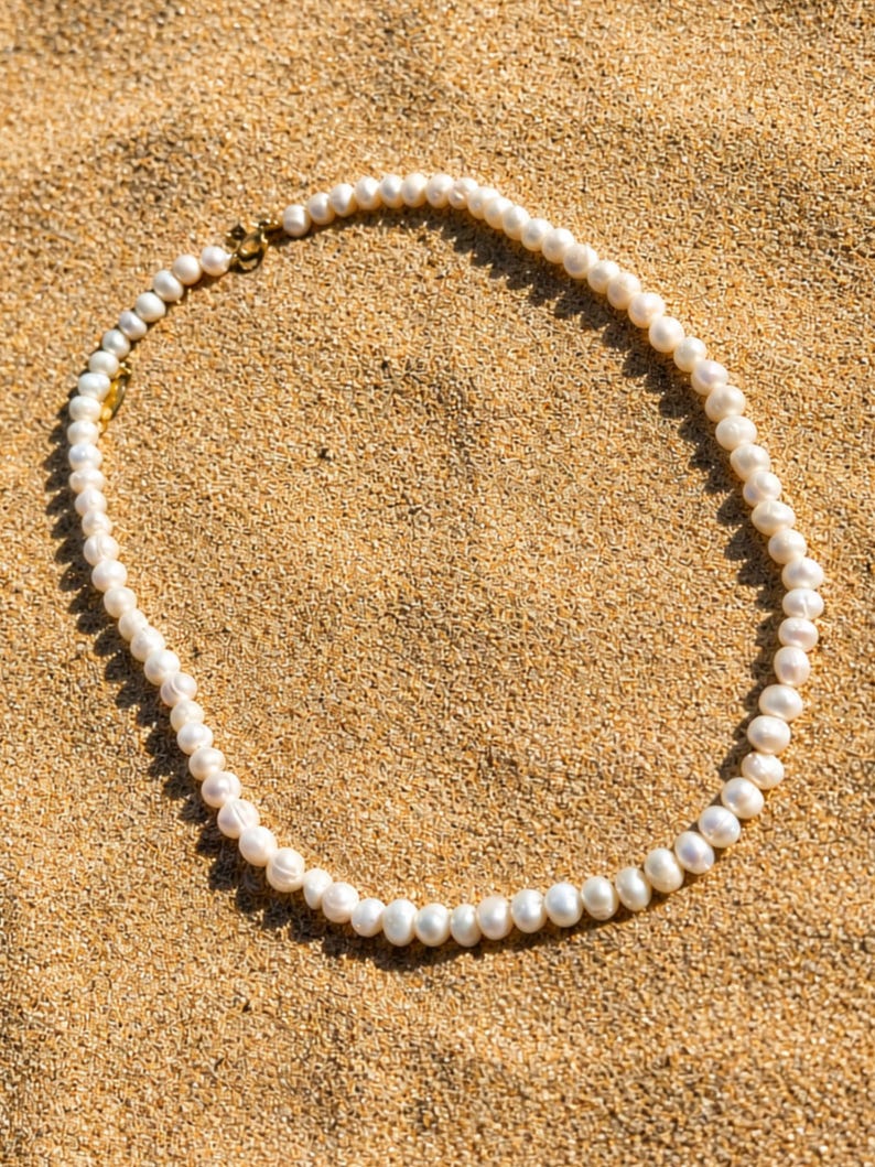 May include: A delicate pearl necklace with a gold clasp rests on a bed of sand. The necklace features numerous white, irregularly shaped pearls strung together. The gold clasp adds a touch of elegance to the natural beauty of the pearls.